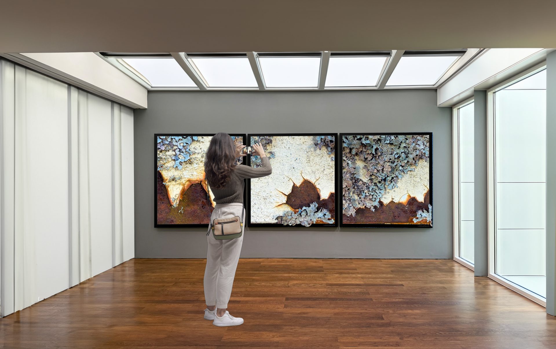 Besucherin fotografiert dreiteilige abstrakte Wandbilder in einer lichtdurchfluteten Kunstgalerie mit Holzfußboden